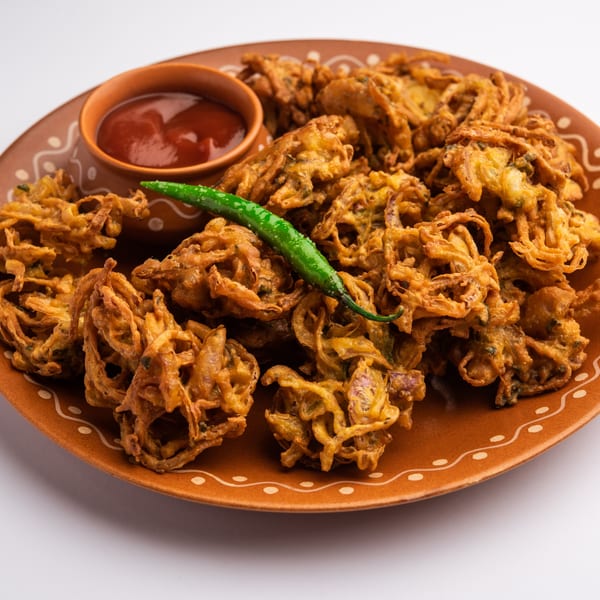 Crispy Indian Wok onion pakoras with a side of red sauce and a green chili on a brown plate.