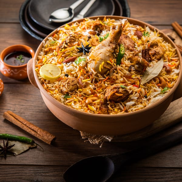 A large bowl of Indian Wok's chicken biryani with spices, lemon, and sauce on a wooden table.